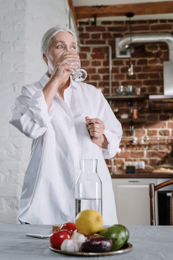 Senior woman drinking water in a cozy kitchen setting with fresh vegetables on the table.